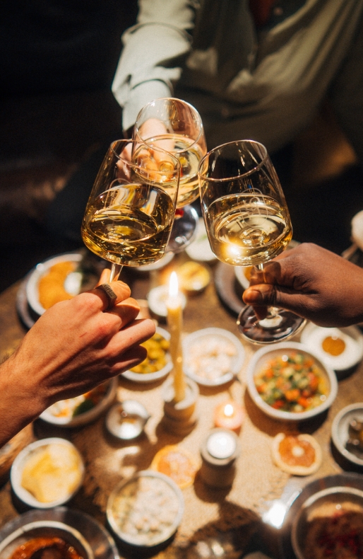 Drei Hände stoßen mit Weingläsern an, die mit Weißwein gefüllt sind, über einem reich gedeckten Tisch mit Mezze und einer brennenden Kerze im Kerzenständer.