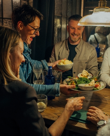 Vier lächelnde Freunde sitzen an einem Holztisch, wobei zwei Personen eine große Platte mit Salat und eine Schale mit Kartoffelchips an den Tisch reichen.