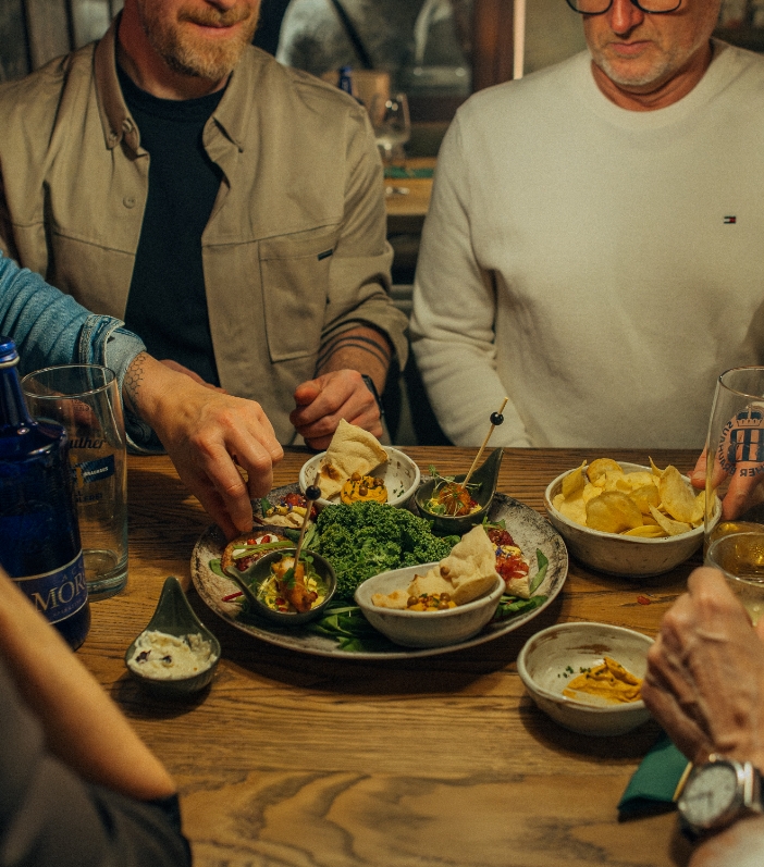 Gruppe von Freunden am Holztisch im Restaurant, die gemeinsam von einer großen Vorspeisenplatte mit Dips, Brot und Salat essen; dazu Chips und Getränke.