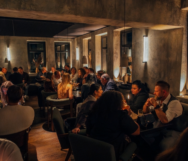 Blick in einen gut besuchten Gastraum am Abend mit Wänden in Betonoptik, warmer Beleuchtung durch Hängeleuchten und vielen Gästen, die an dunklen Tischen sitzen und sich unterhalten.