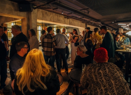 Belebte Szene in einem Restaurant mit vielen stehenden Gästen, die sich unterhalten und trinken; im Hintergrund eine beleuchtete Bar und eine Decke im Industrie-Look.