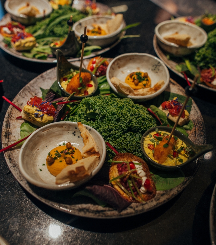 Nahaufnahme einer rustikalen Vorspeisenplatte mit frischem Grünkohl in der Mitte, umgeben von Hummus-Schälchen mit Pita-Brot, Falafel-Bällchen in Sauce und kleinen Canapés.