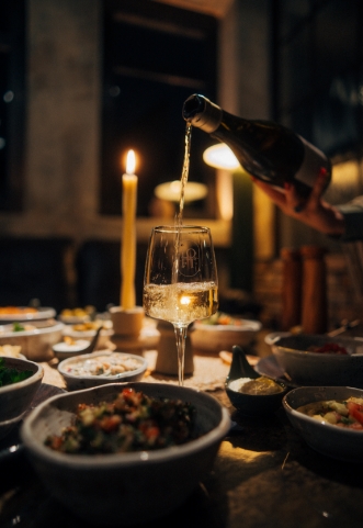 Hand schenkt Weißwein aus einer Flasche in ein stilvolles Weinglas auf einem reich gedeckten Tisch, umgeben von Schalen mit verschiedenen Speisen und einer brennenden Kerze im Hintergrund.